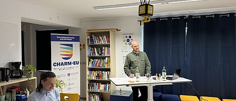 There are two people in the picture. One person is presenting something and the other is writing on a laptop. In the background is a banner with the inscription ‘CHARM-EU’.