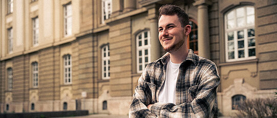 Tim studiert Diversitätsmanagement an der JMU. Er steht vor dem Gebäude der Universität Würzburg am Wittelsbacherplatz.