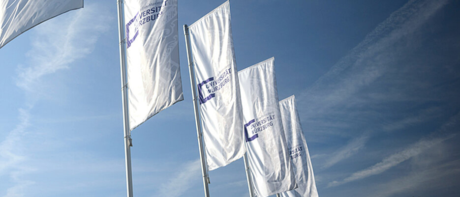 Wehende Flaggen mit dem Logo der Uni Würzburg vor blauem Himmel.